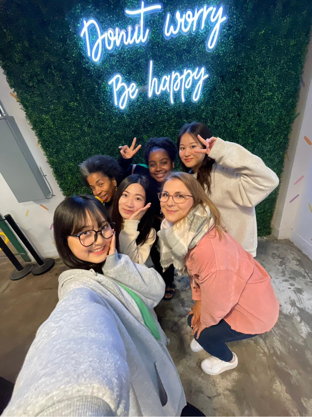 A group of six young women are happily posing in front of a green wall featuring a neon sign that reads, "Donut worry, Be happy".