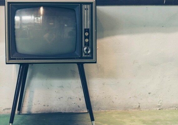 An old CRT television set is displayed on a tripod stand, positioned against a textured wall.