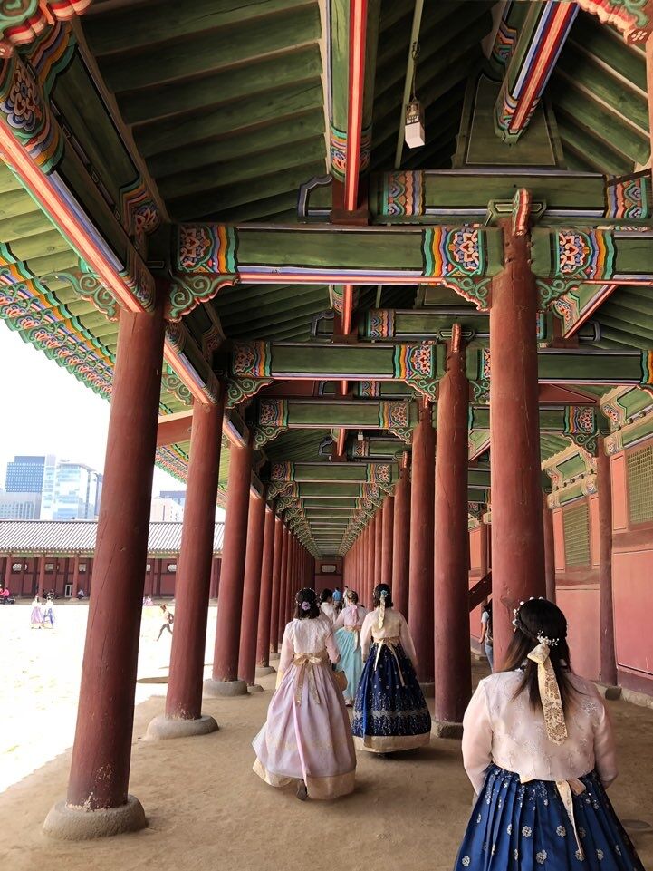 Three women in traditional Korean clothing called Hanbok walk through a covered walkway featuring red pillars and ornately painted ceilings.