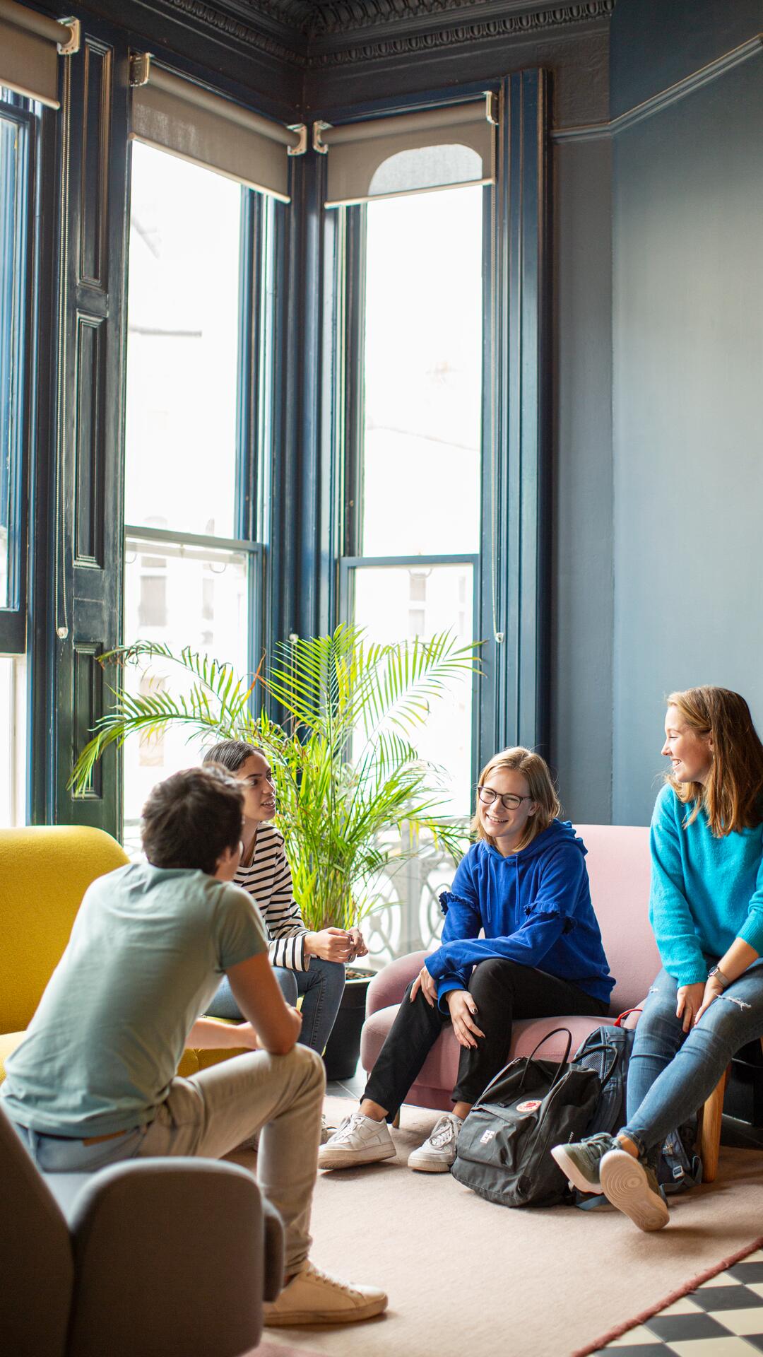 EF Language Abroad students relaxing and talking together in a bright common room, sitting on colorful sofas near tall windows and plants.