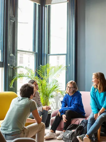 EF Language Abroad students relaxing and talking together in a bright common room, sitting on colorful sofas near tall windows and plants.