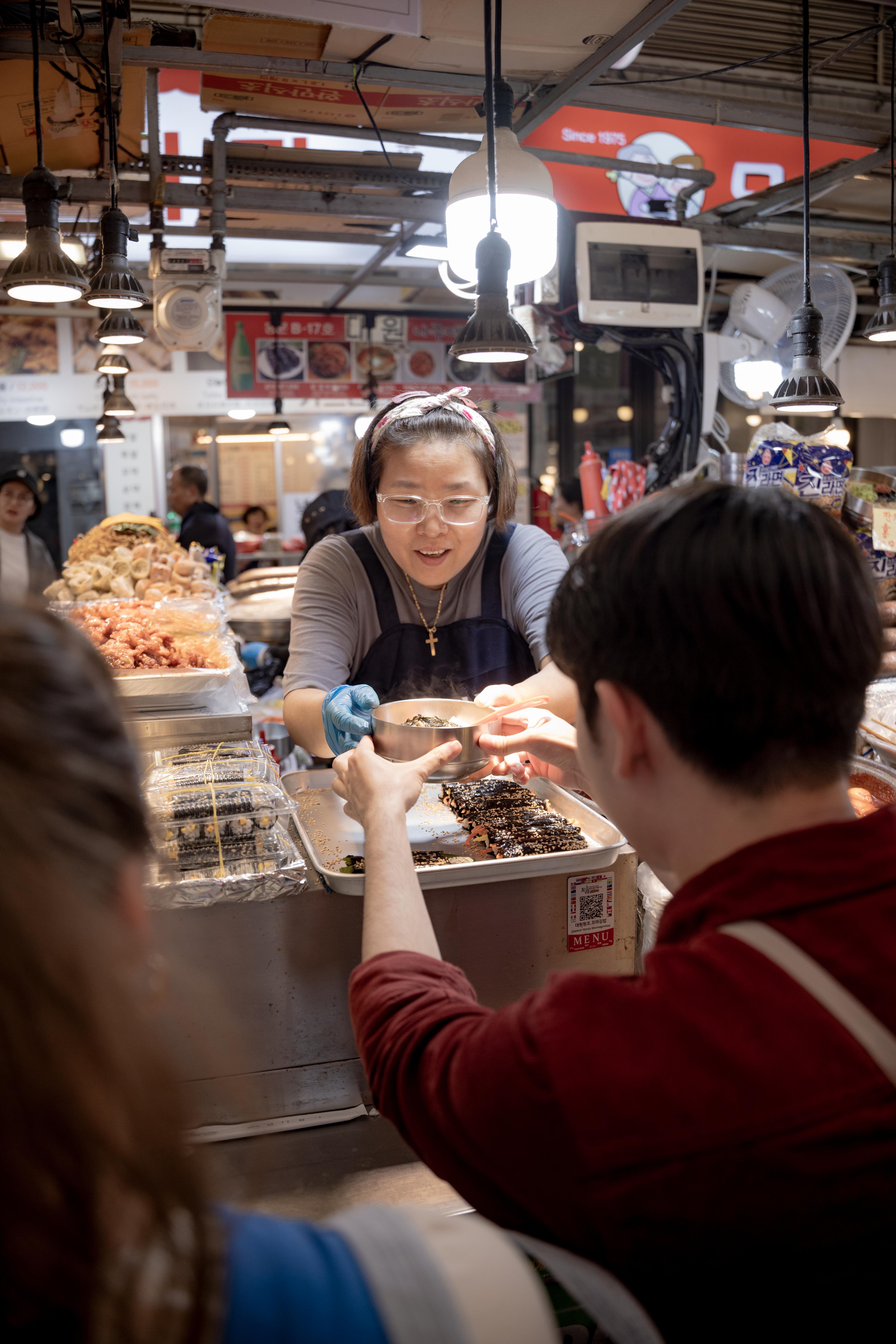 Vendor serving food at a busy market as students explore nearby stalls