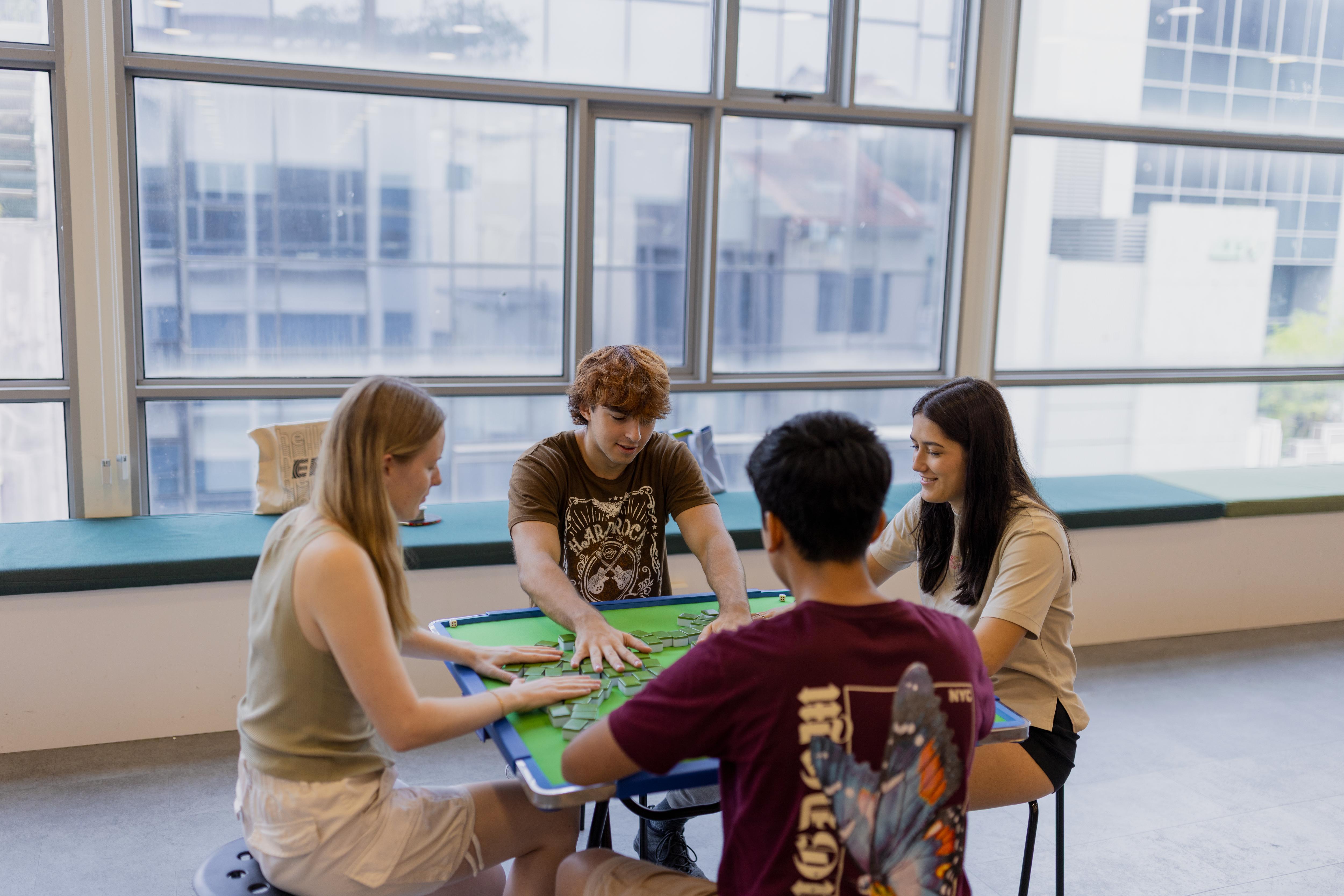 Four students enjoy a friendly game of Mahjong, socializing and connecting in a bright, relaxed campus setting