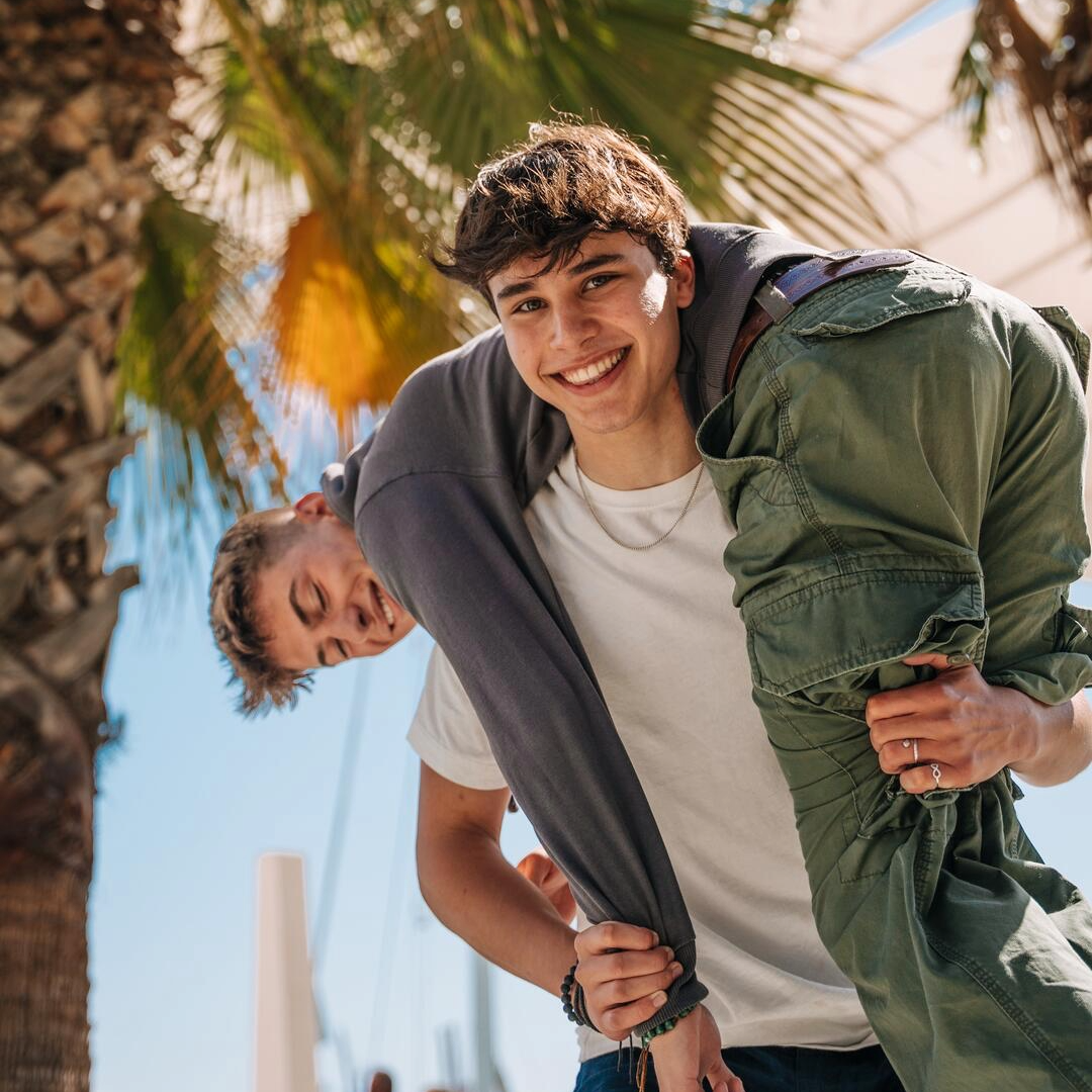 Two EF Language Abroad students having fun outdoors in Málaga, Spain, with one playfully carrying the other over his shoulder in a sunny waterfront area lined with palm trees.
