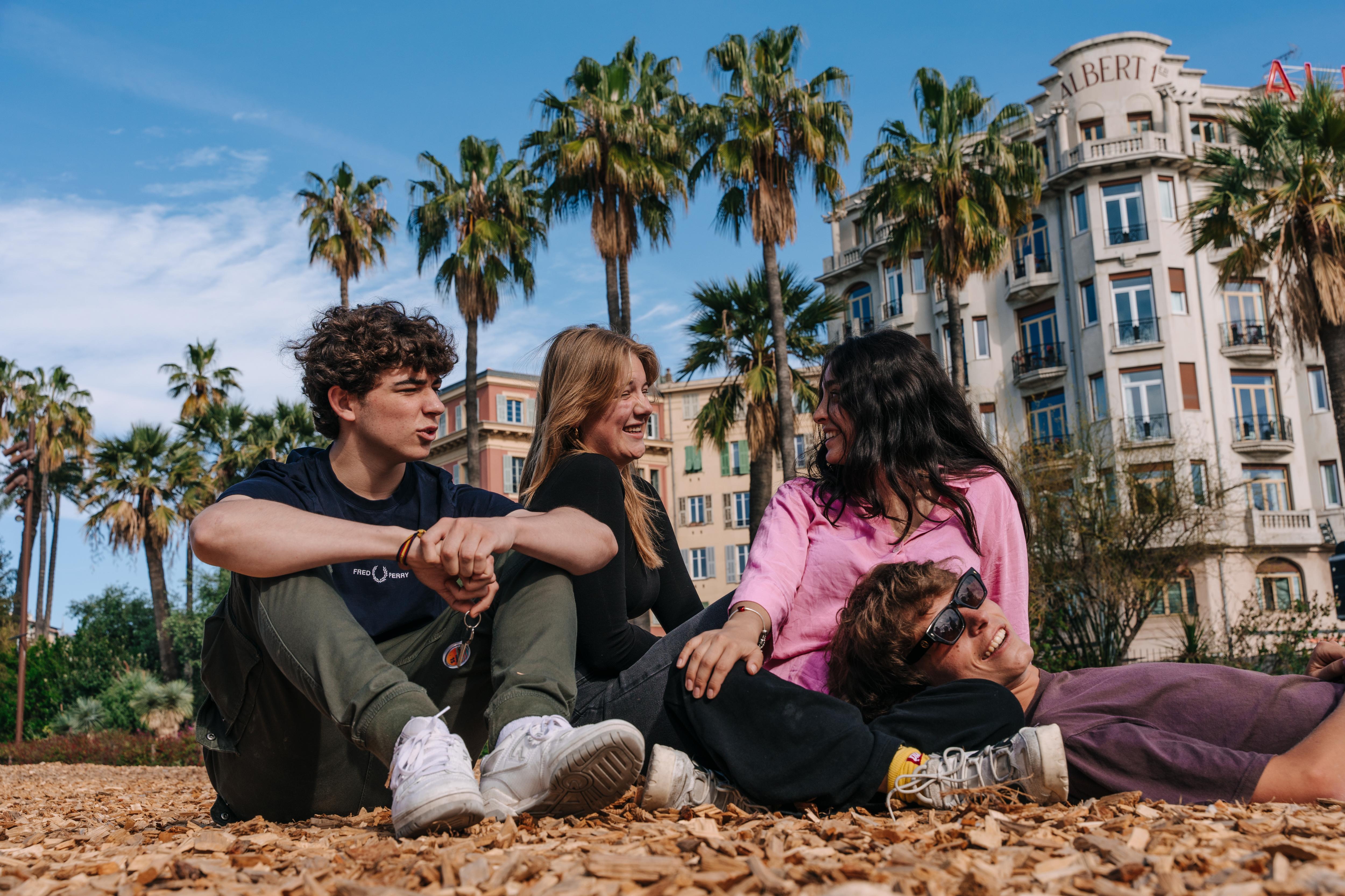 EF Language Abroad students relaxing and laughing together in a sunny park in Nice, France, sitting on the ground under palm trees with historic buildings in the background.