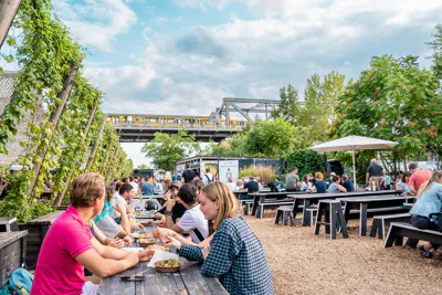 EF Language Abroad students eating and talking together at an outdoor beer garden in Berlin, with long picnic tables, greenery, and an elevated yellow U-Bahn train passing in the background.