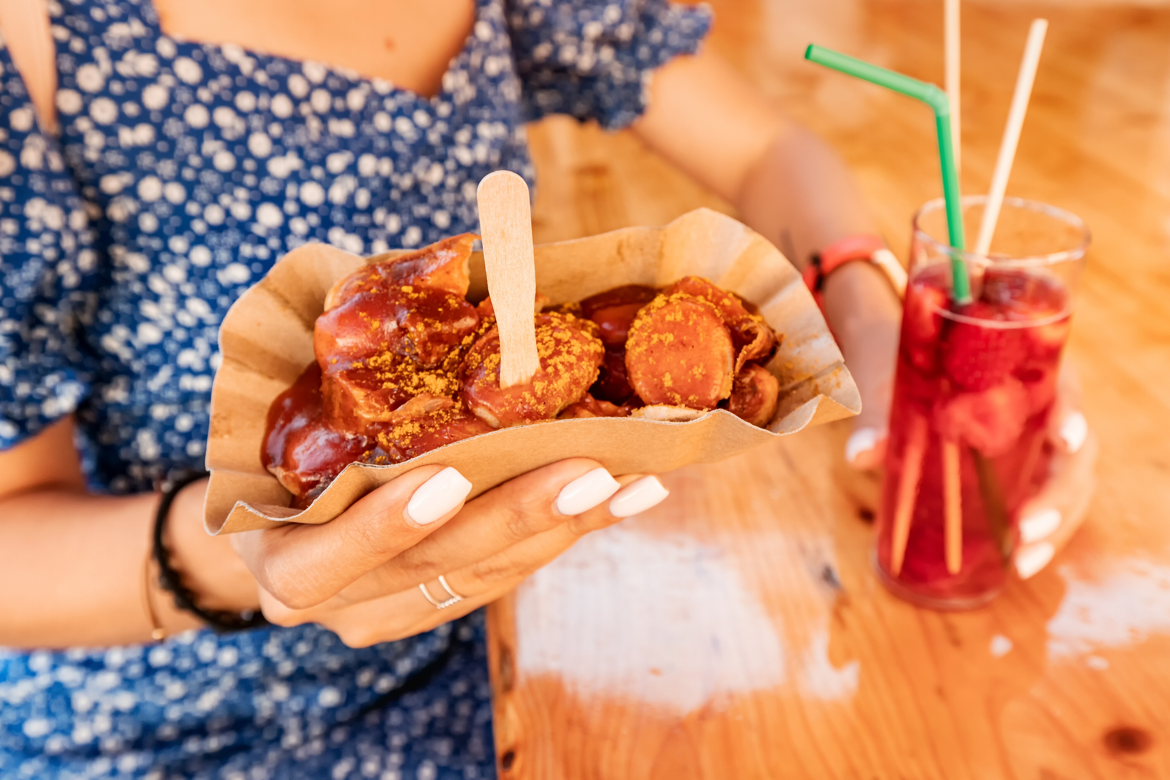 A person is holding a serving of currywurst with fries and a drink. The food is served in a cardboard container and garnished with a fork