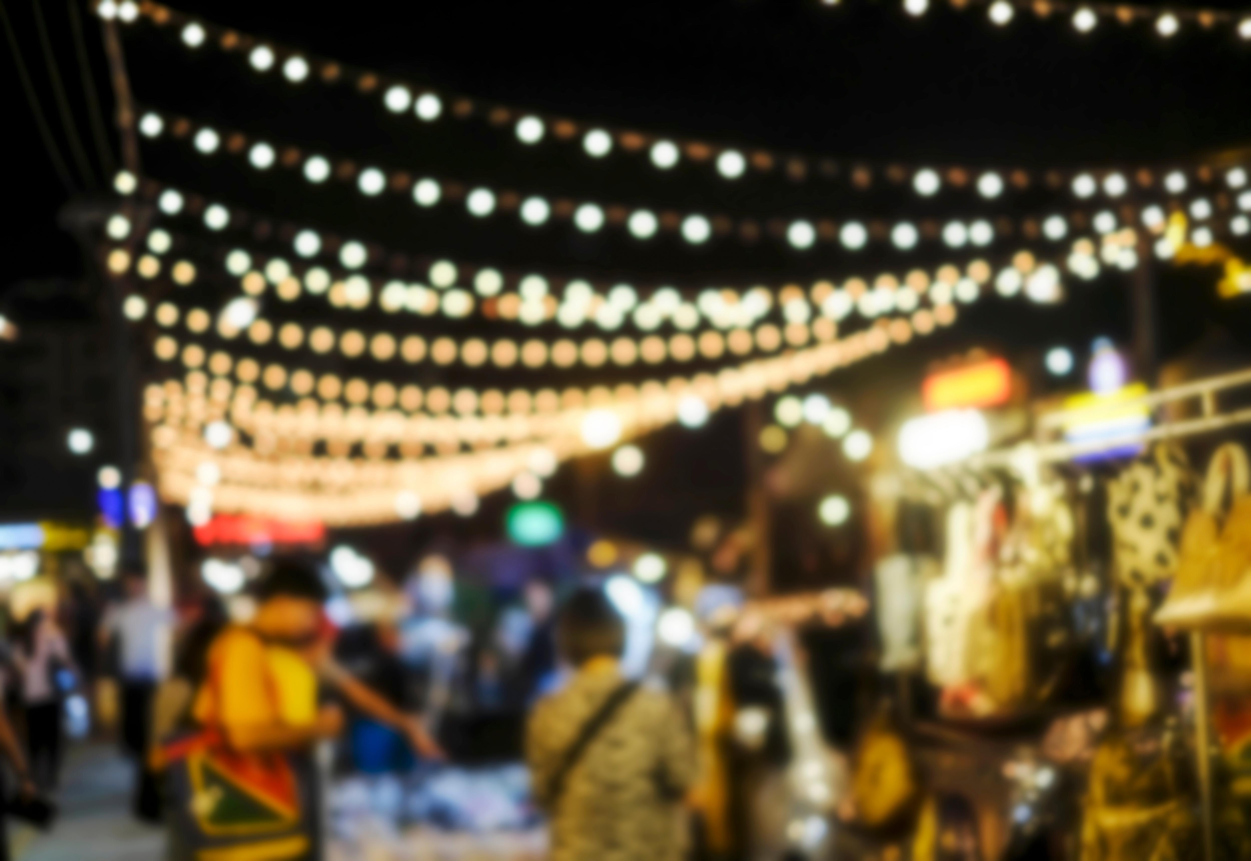 A blurry night shot of a bustling market, with strings of lights draped overhead, illuminating crowds of people and various stalls