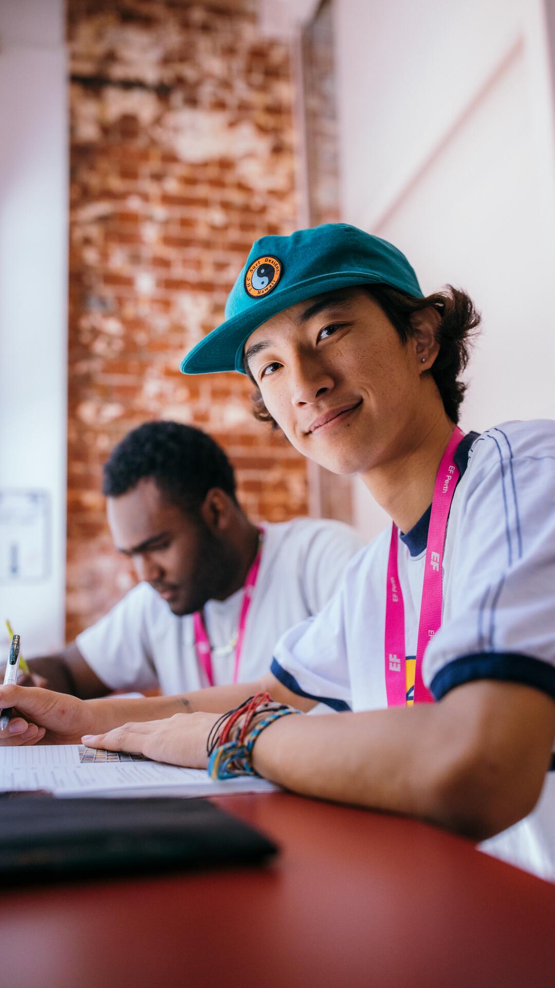 EF Language Abroad students studying together in a classroom, with one student smiling toward the camera while working on an assignment at their desk.