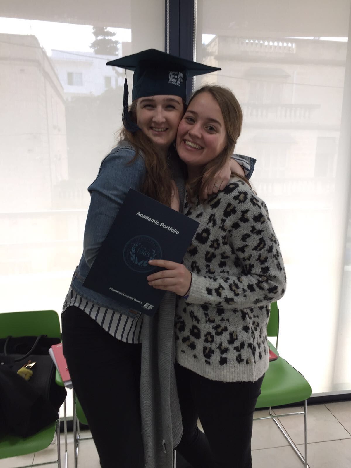Two young women are hugging, one is wearing a graduation cap and holding a certificate. They appear to be celebrating their graduation.