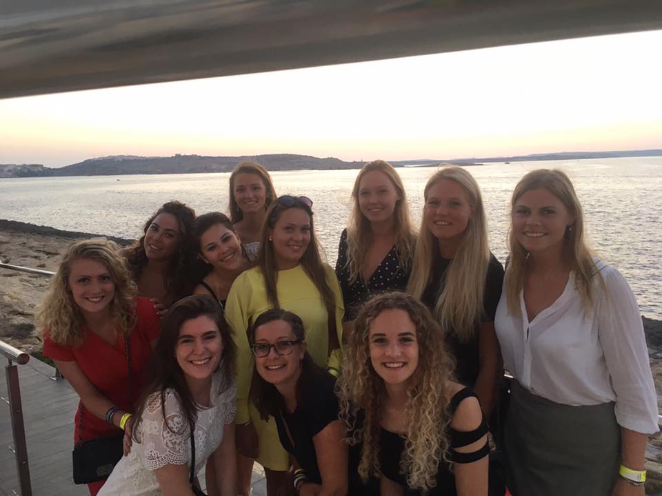 A group of eleven young women are posing for a photo on a warm evening, with a body of water and a distant landmass visible in the background.