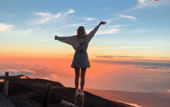 A person stands with arms outstretched, embracing a breathtaking sunset over a sea of clouds. The warm, golden light illuminates the sky, creating a serene and awe-inspiring moment of natural beauty.