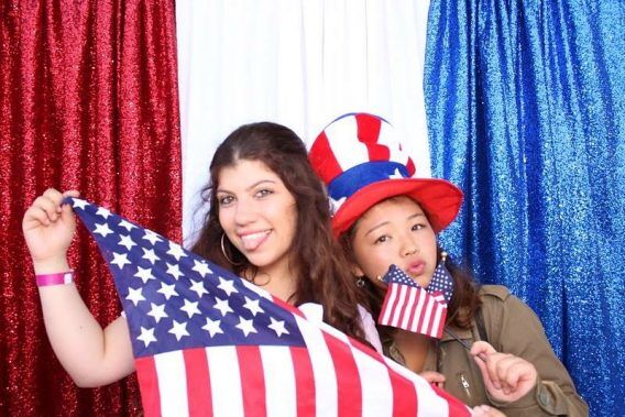 Two young women are playfully posing for a photo with an American flag and a patriotic hat, in front of a sparkling red, white, and blue backdrop.