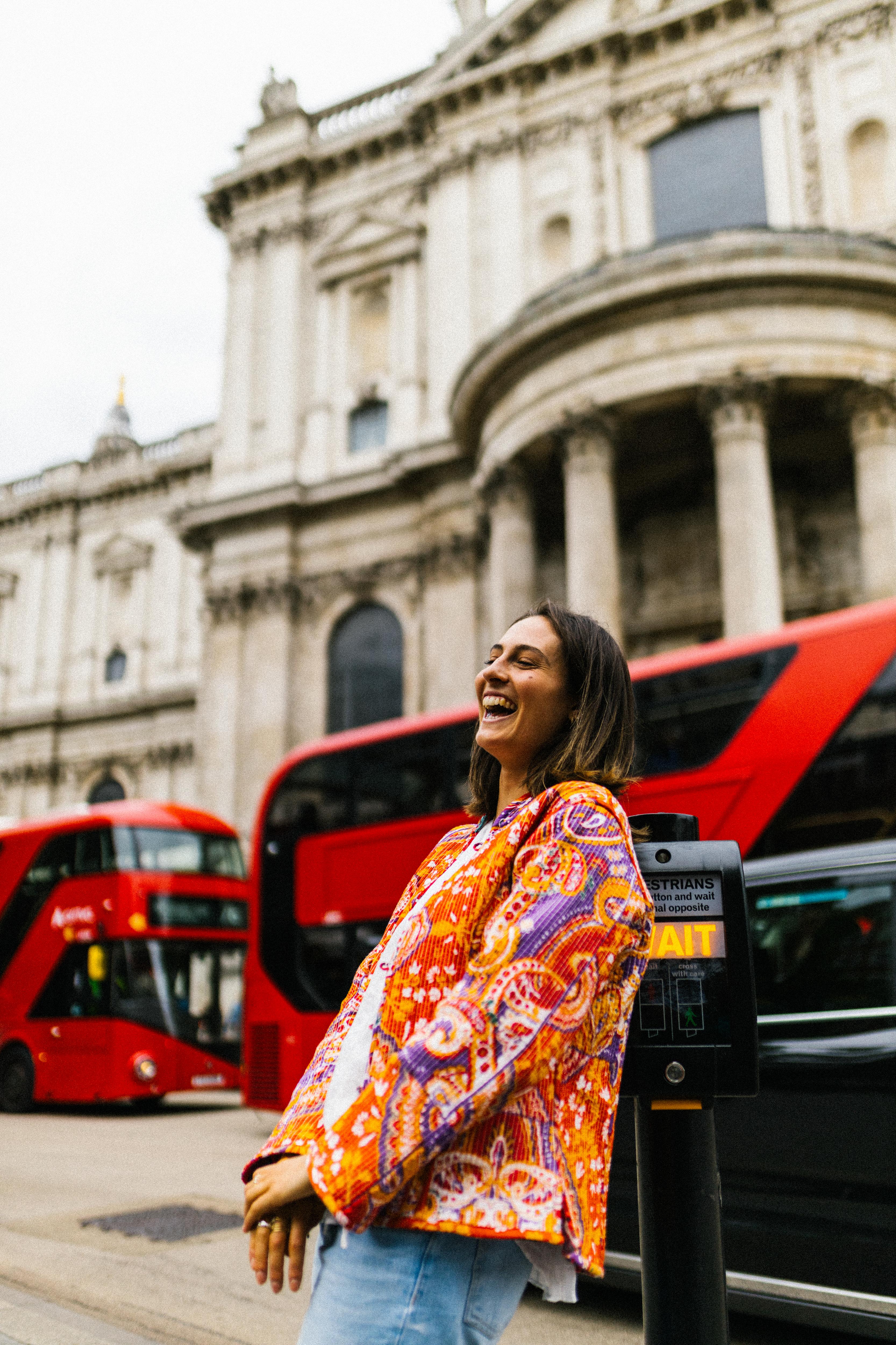 A woman laughs in front of a red double-decker bus and a historic building in London.