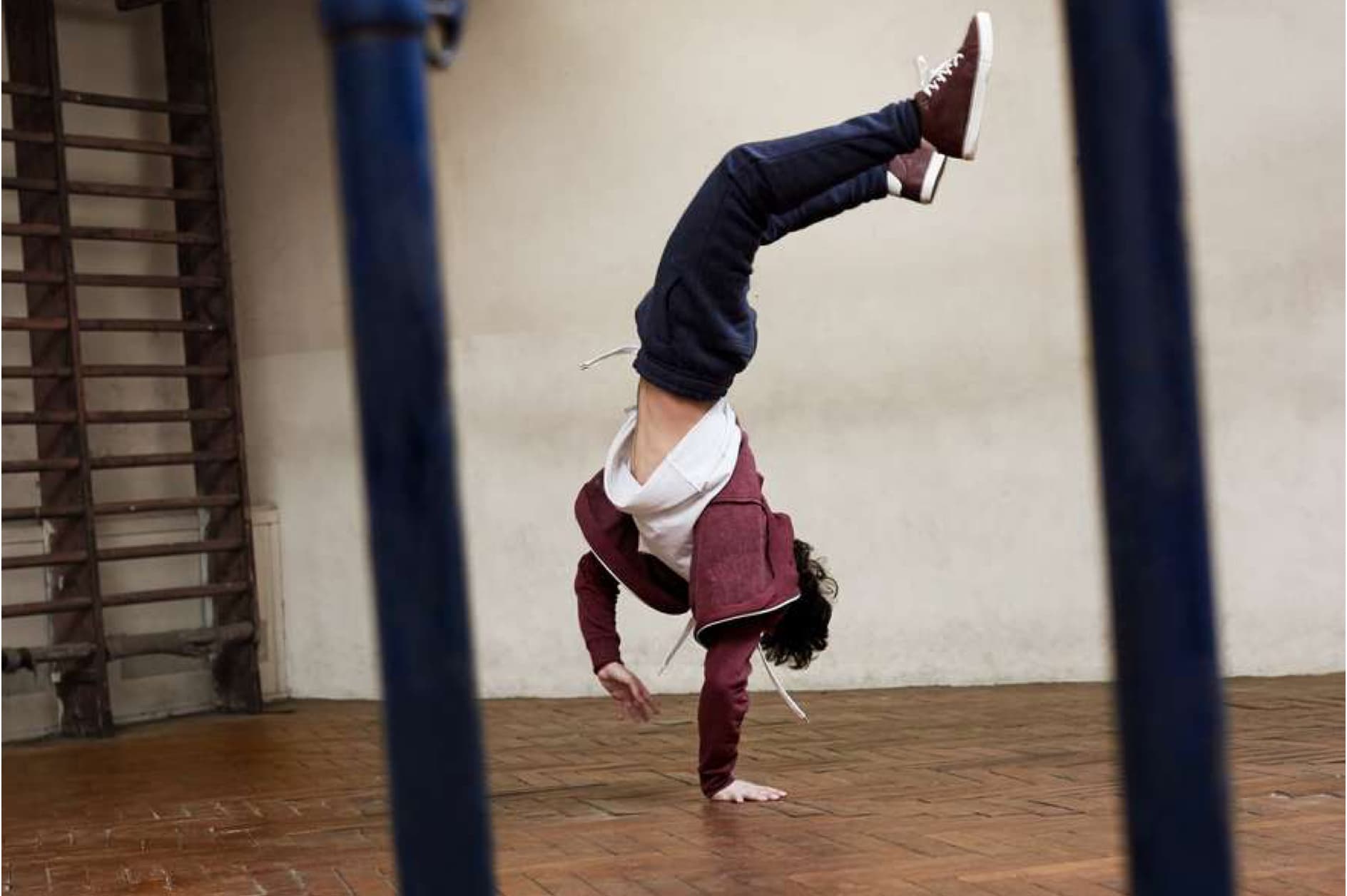 A person is captured mid-action performing a handstand on a wooden floor. Their legs are extended upwards, showcasing their athletic ability and balance. The individual is dressed in casual athletic wear.