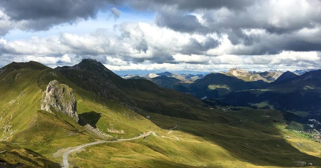 A winding road snakes through lush green mountains under a dramatic, cloudy sky, with shadows and sunlight creating a dynamic landscape.