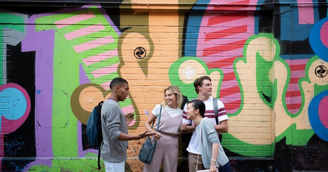 Four young adults are standing in front of a vibrant graffiti wall. They appear to be engaged in a casual conversation, enjoying a moment together.