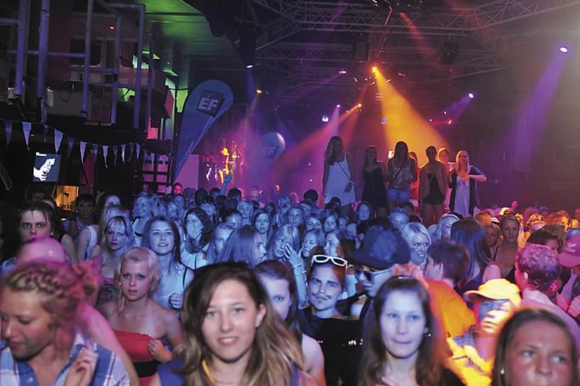 A large crowd of people are dancing and enjoying themselves in a dark venue, illuminated by colorful lights and stage fog.