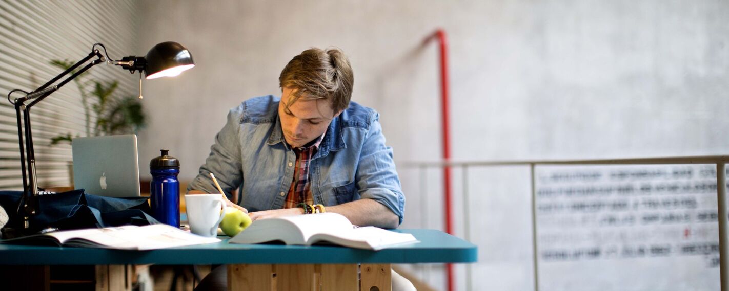 A young man sits at a desk, writing in an open book, illuminated by a desk lamp. A laptop, water bottle, and apple are on the desk.
