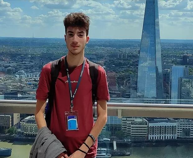 A young man with dark, curly hair stands in front of a cityscape featuring the Shard building. He is wearing a dark red t-shirt and has a backpack on.