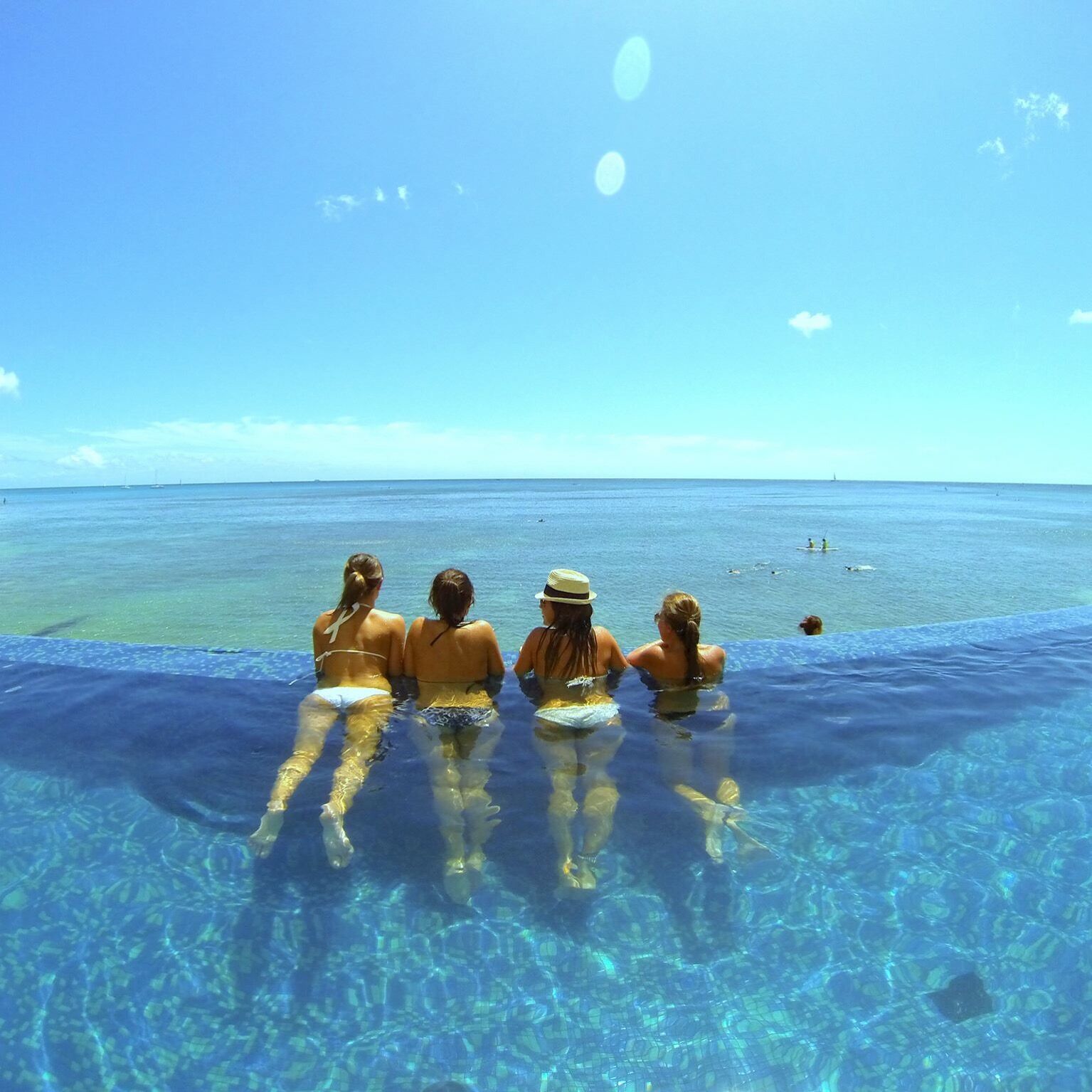 Four women are enjoying a sunny day in an infinity pool, overlooking the turquoise ocean and blue sky.