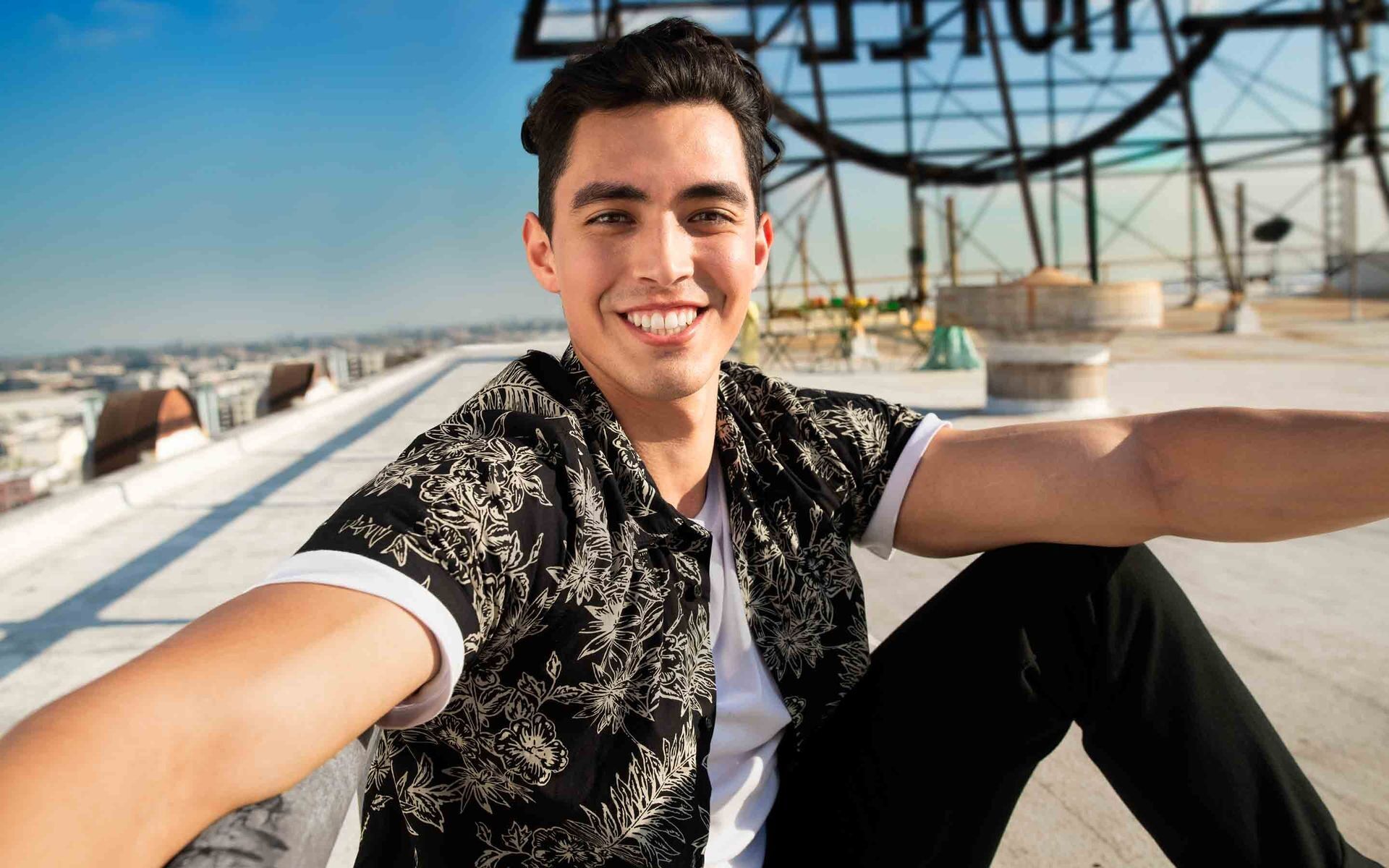 A young man with dark hair and a bright smile is taking a selfie on a rooftop, with the iconic Hollywood sign visible in the background under a clear blue sky.