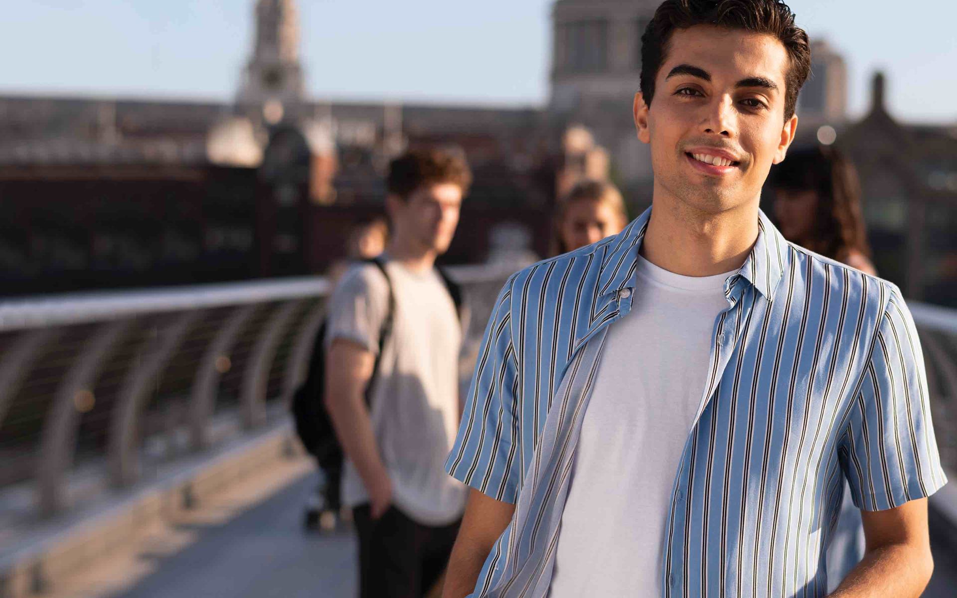 A young man with dark hair, wearing a blue striped shirt over a white t-shirt, walks across a bridge with other people visible in the background.