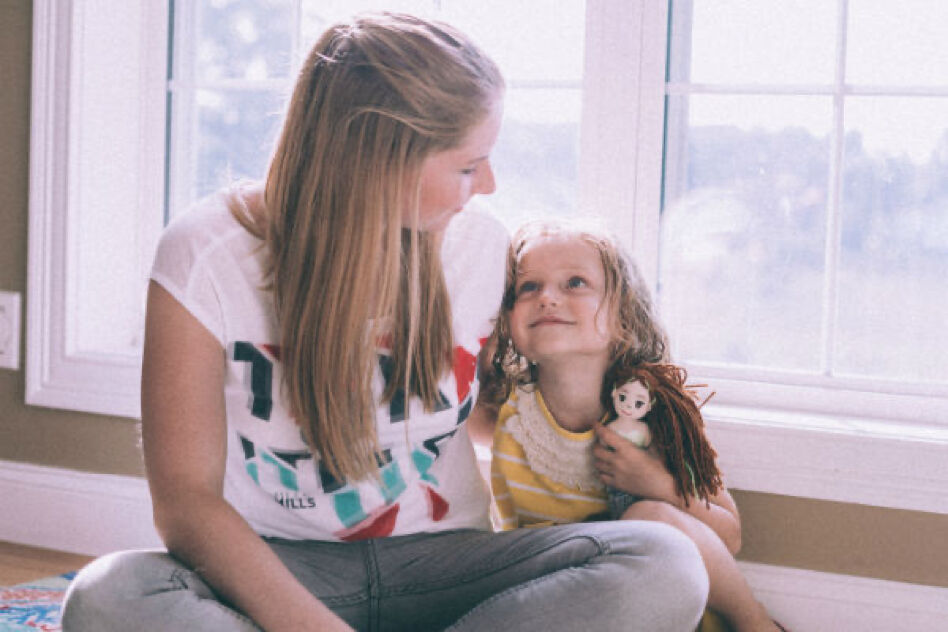 Two young girls are sitting on the floor in front of a window. The older girl is holding the younger one, who is holding a doll.