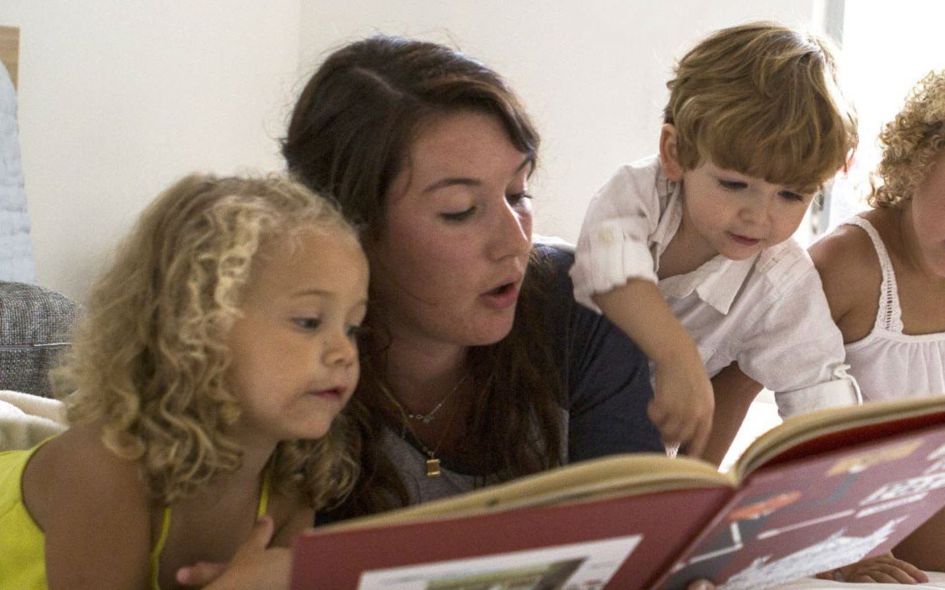 A young woman is attentively reading a book to three small children, who are watching with rapt attention.