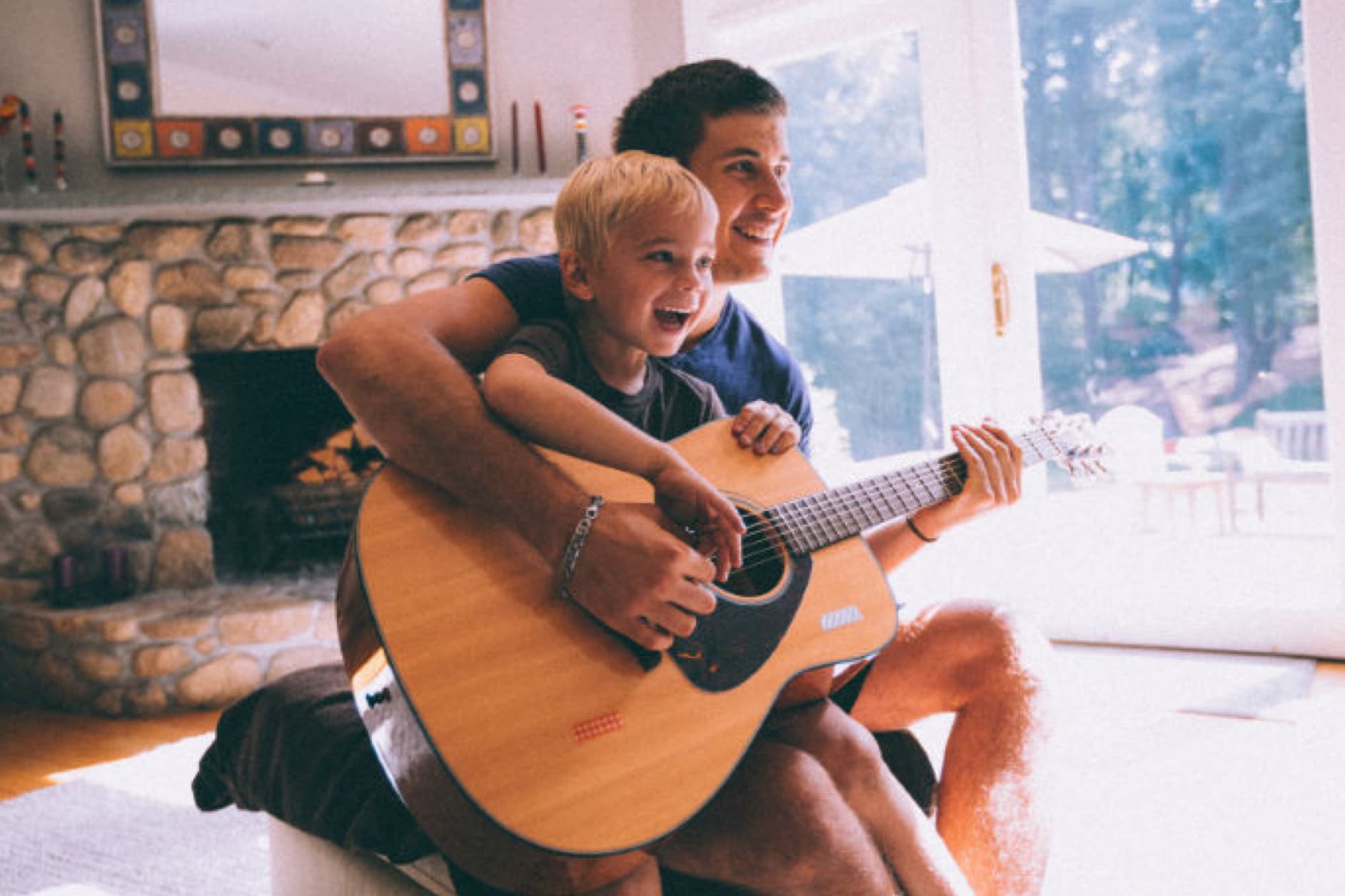 A man and a child are sitting on a lap playing the guitar. They are smiling and appear to be having a lovely time together.