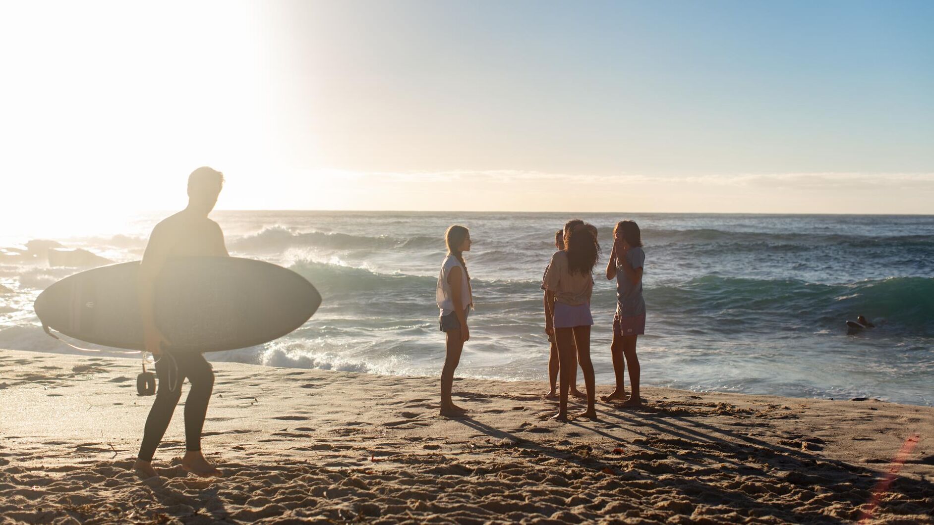 A group of friends enjoys a sunny day at the beach as a surfer looks out at the ocean with his surfboard. The waves are gentle and the atmosphere is relaxed.
