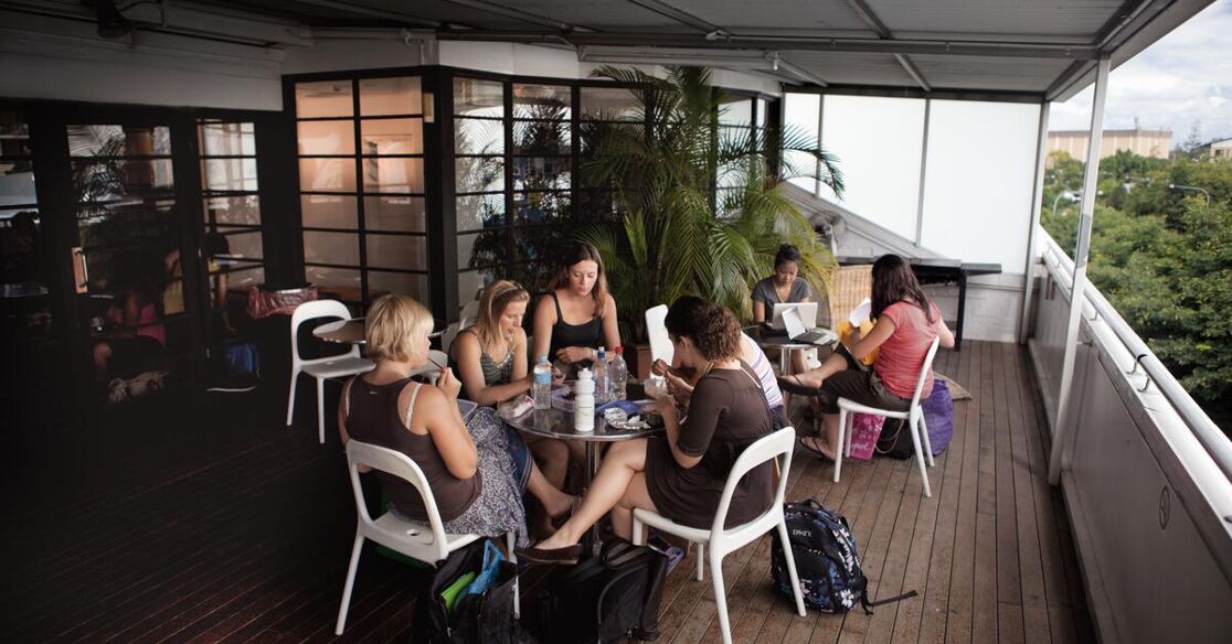 A group of women are gathered around a circular table on a balcony, engaged in conversation, with trees visible in the background.