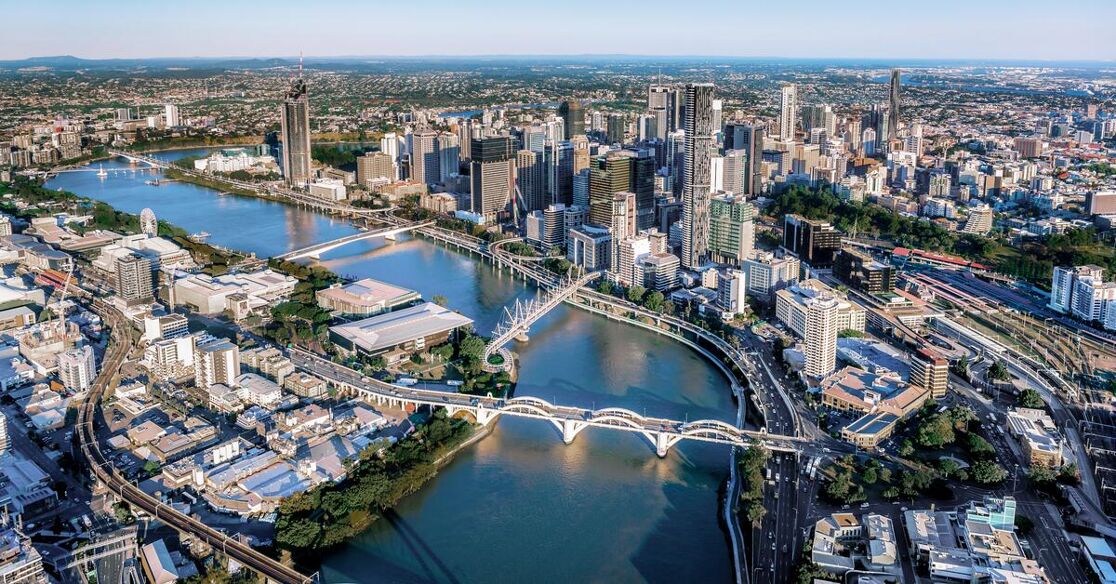This is a wide aerial view of a bustling city with a river winding through it, crossed by multiple bridges. Tall buildings line the riverbanks, creating an impressive skyline.