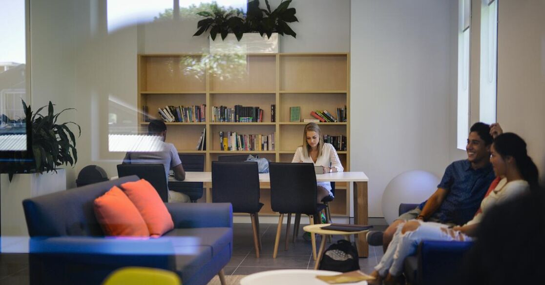 A group of students are studying and relaxing in a modern library or common area, featuring comfortable seating and numerous books.