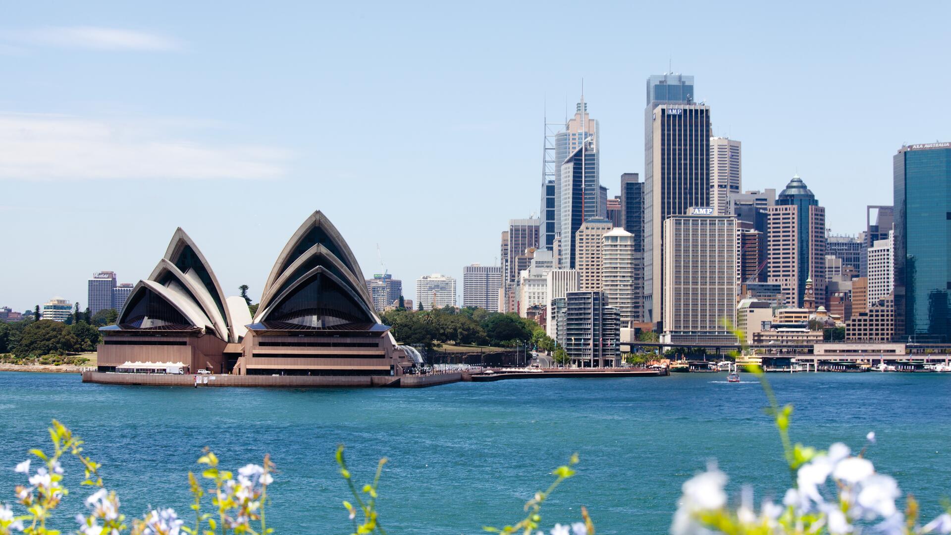 The image shows a panoramic view of the Sydney Opera House and the city skyline, with a body of water in the foreground and lush greenery on the left.