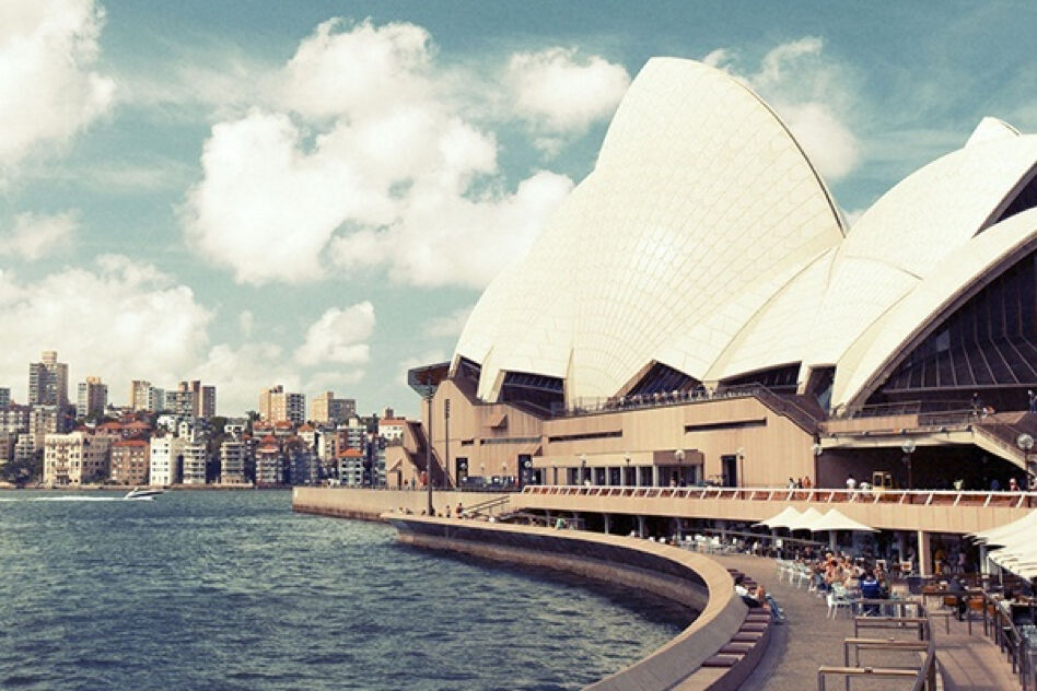 This image showcases the iconic Sydney Opera House in Australia, featuring its distinctive sail-like architecture rising above the water. The city's skyline is visible in the background.
