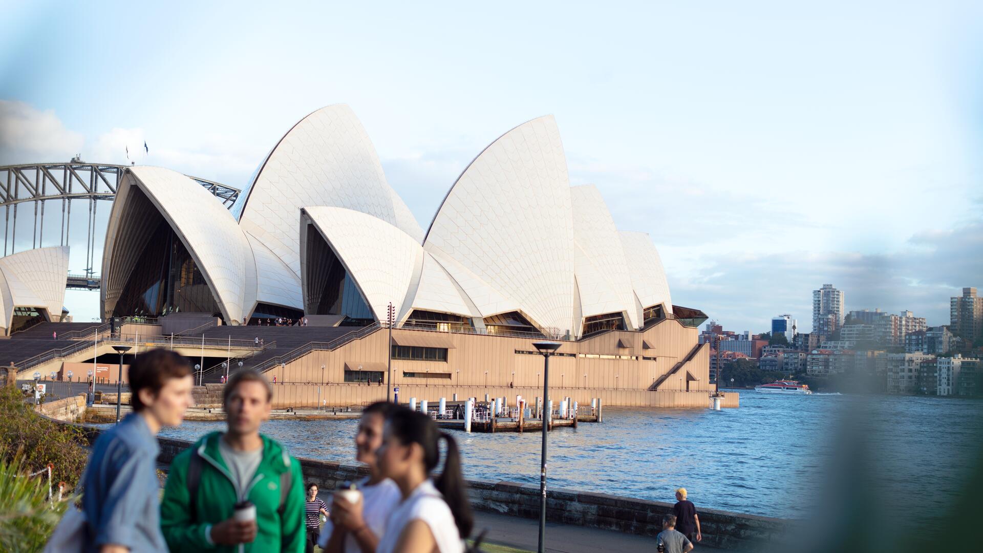 Several people are walking along the waterfront on a sunny day, with the Sydney Opera House and Harbour Bridge visible in the background.