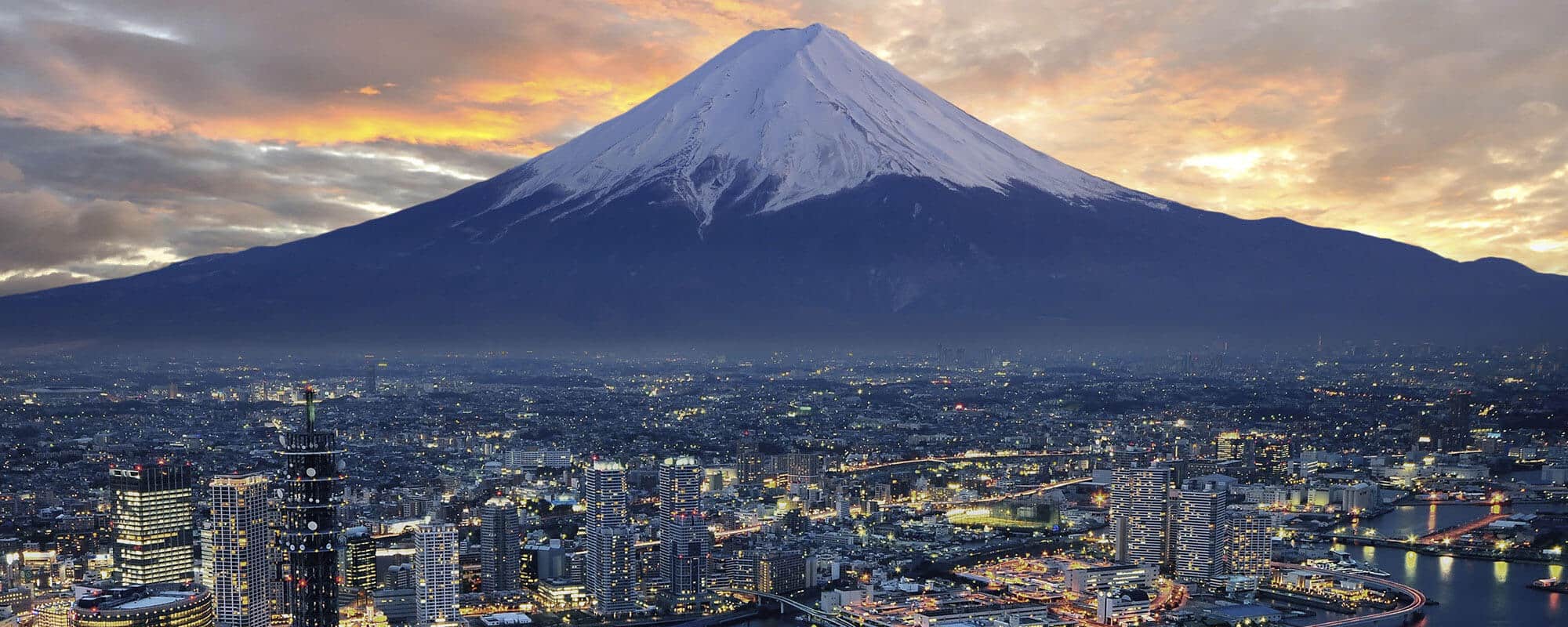 Mount Fuji, an active volcano and Japan's tallest peak, stands majestically over a lit-up city at sunset.