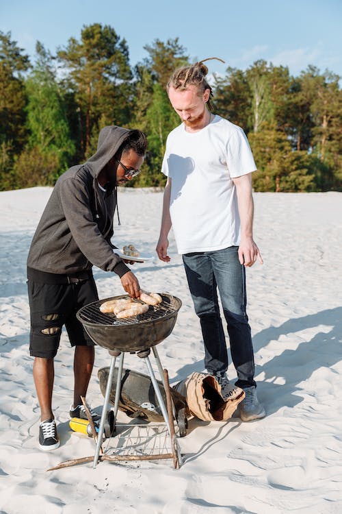 Two men are enjoying a barbecue on a sandy beach, with a forest in the background. They are grilling sausages over a charcoal grill on a sunny day.
