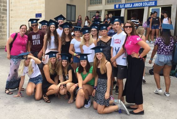 A group of smiling young adults, likely students, are posing for a photo outdoors, all wearing graduation caps. The building behind them has a sign that reads "G.F. Abela Junior College".