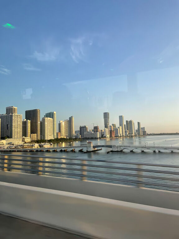 The Miami skyline at sunset, featuring tall buildings rising above the water with a bridge meandering through the scene.