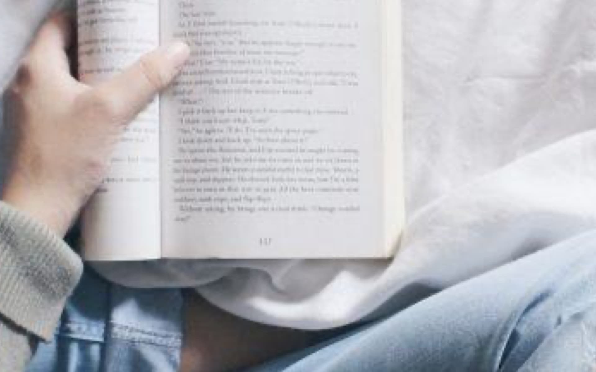 A person is lying in bed, holding an open book and reading. The image has a soft, cozy aesthetic, with a focus on the book and the reader's hands and lap.