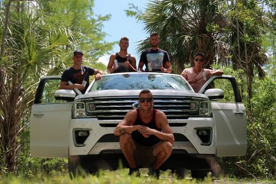 Five young men pose in front of a white SUV, surrounded by lush trees, with one man crouching in the foreground.