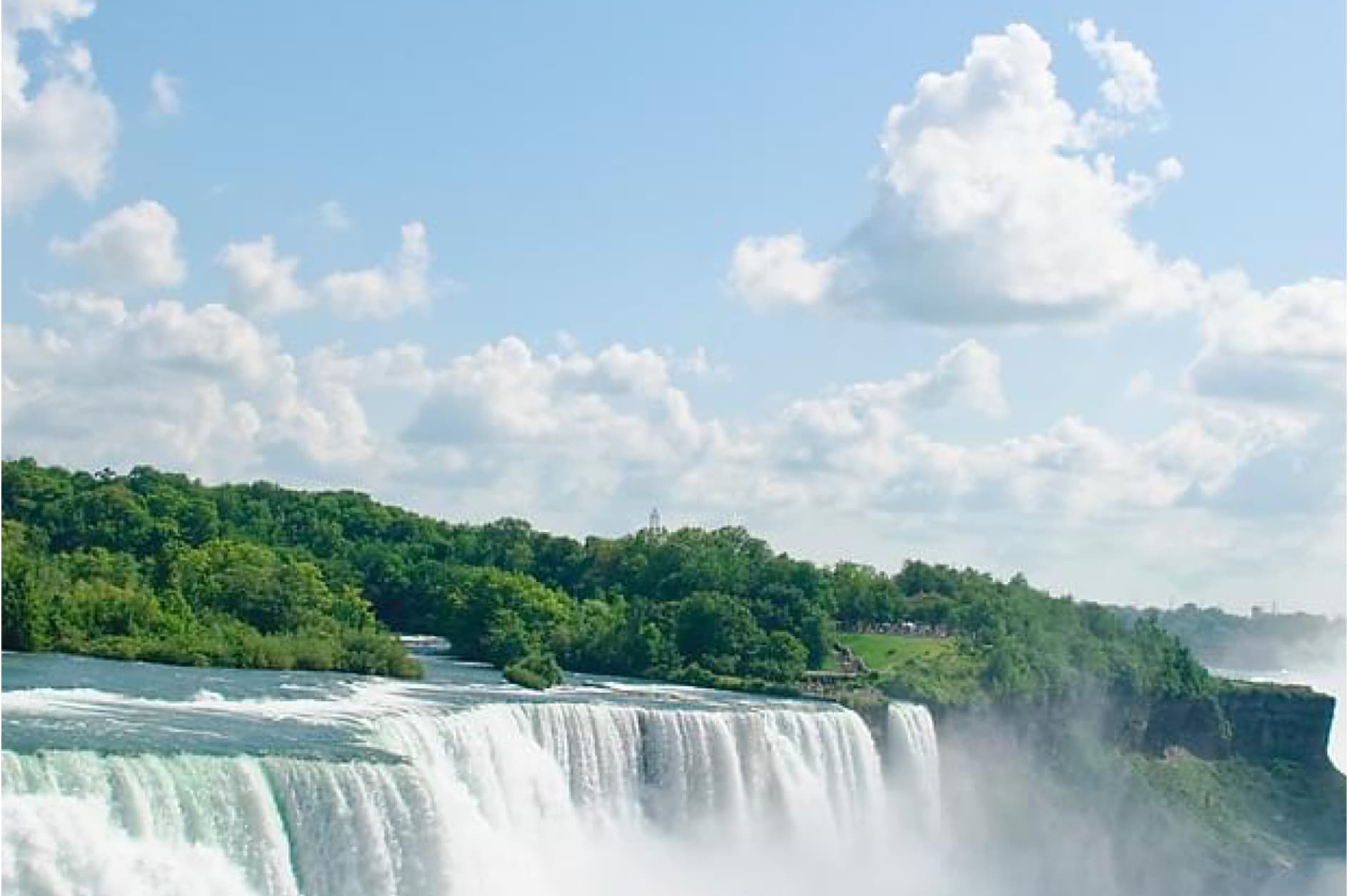 A breathtaking view of the roaring Niagara Falls, framed by lush greenery under a bright blue sky dotted with fluffy white clouds. A truly majestic natural spectacle.
