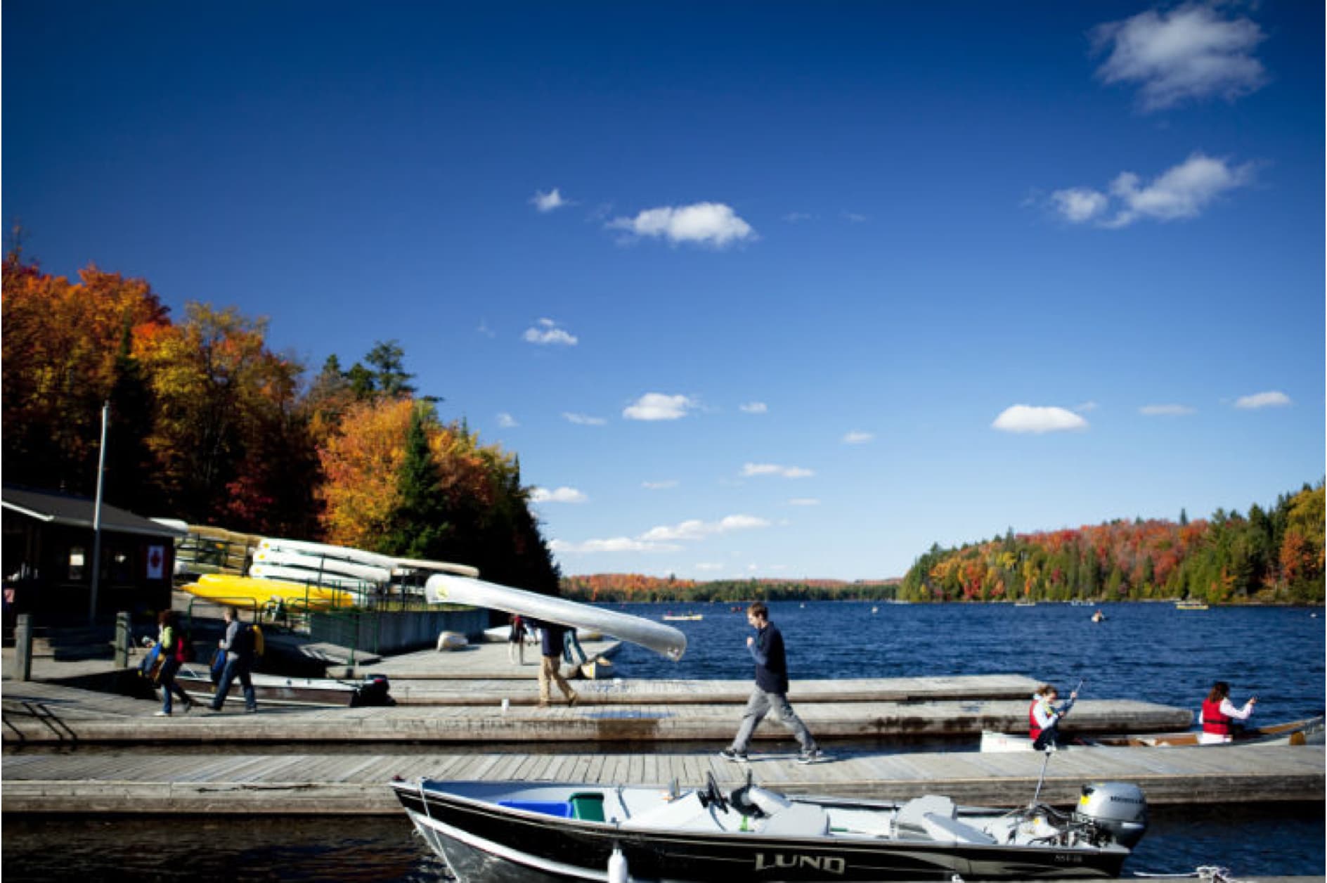 This image depicts a serene autumn day at a lake, with colorful fall foliage framing the water. People are seen near boats and rowing equipment on a wooden dock, suggesting a rowing or boating club.