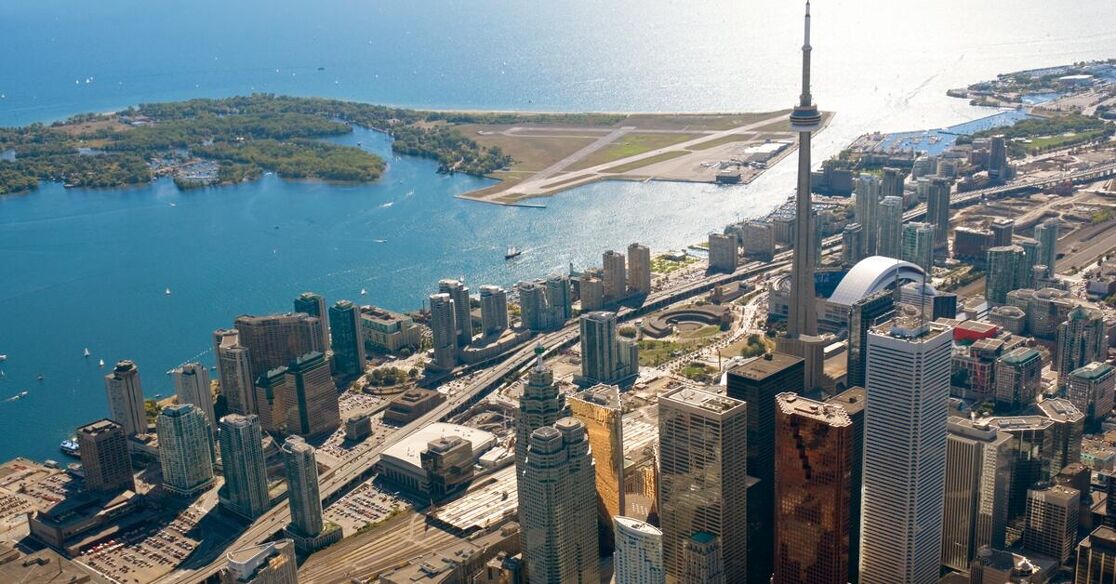 An aerial view of Toronto, Canada, featuring the CN Tower prominently in the foreground, with Lake Ontario in the background. The image showcases the city's skyline and the Toronto Islands.