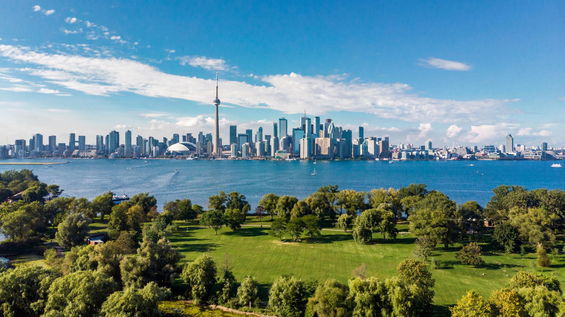 A scenic view of the Toronto skyline, featuring the CN Tower, captured from a park with lush green trees and grass by the shores of Lake Ontario.