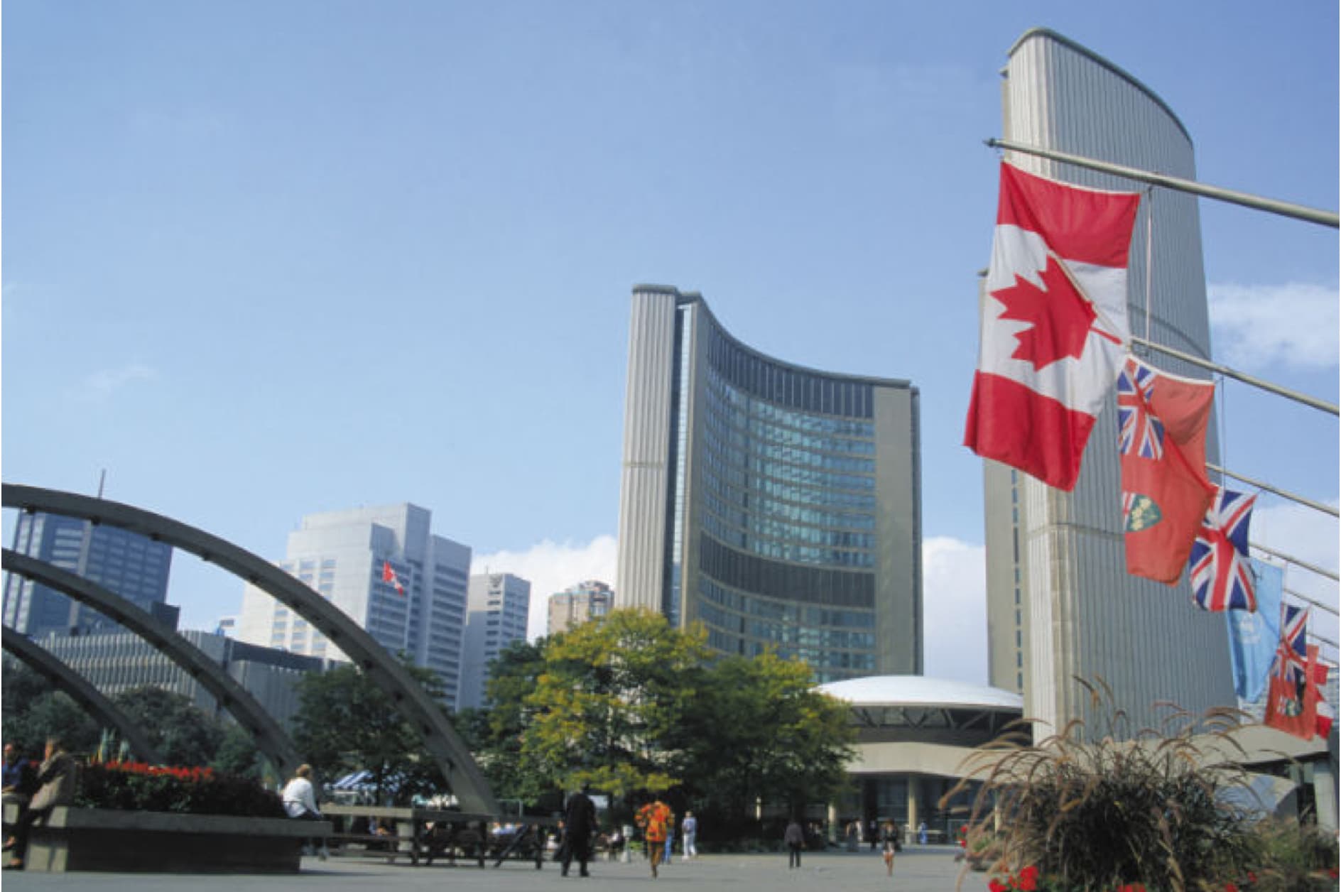 This image shows the Toronto City Hall building, a prominent landmark in the city, with Canadian flags flying in the foreground under a clear blue sky.