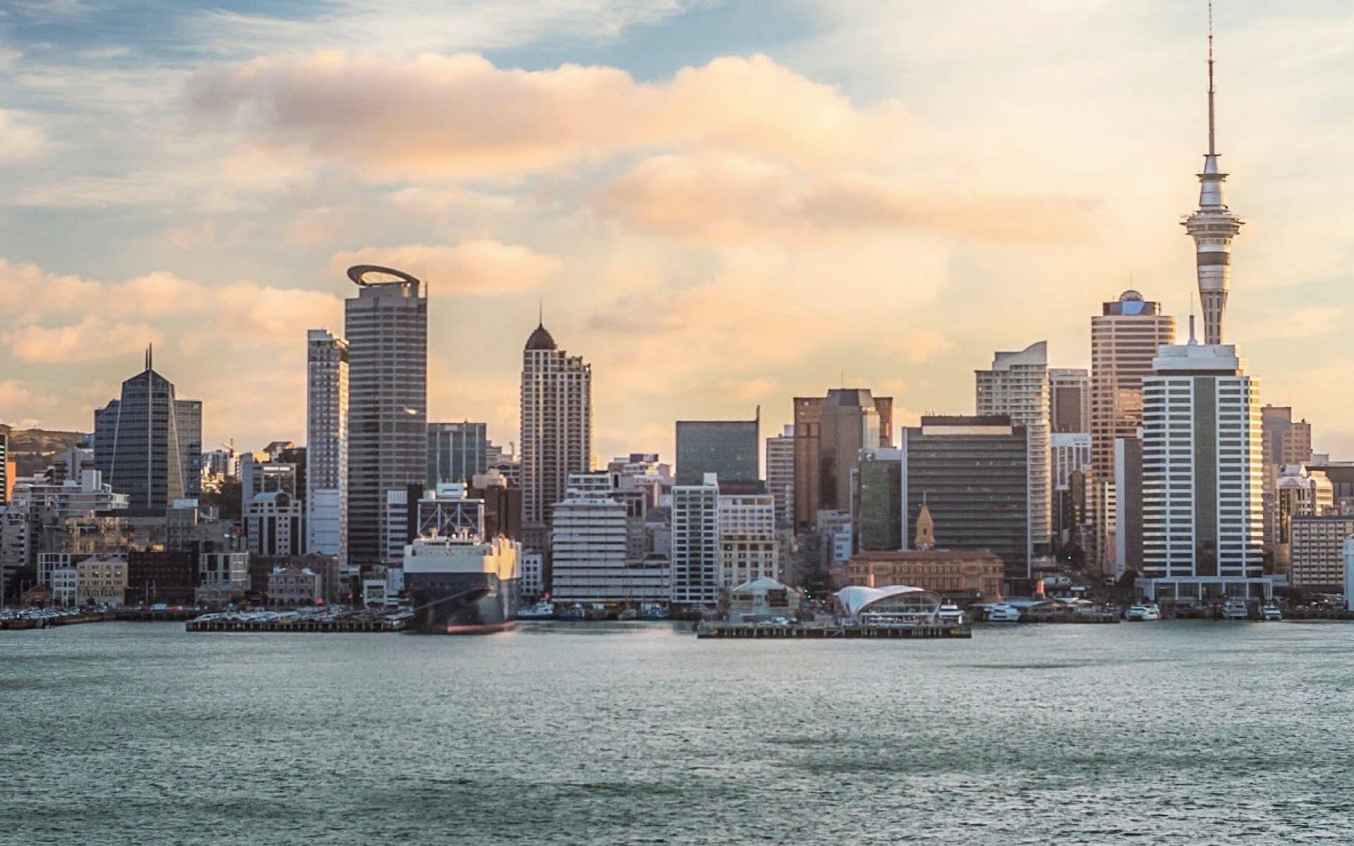The skyline of Auckland, New Zealand, featuring the Sky Tower and modern skyscrapers rising above the water under a dramatic sky.