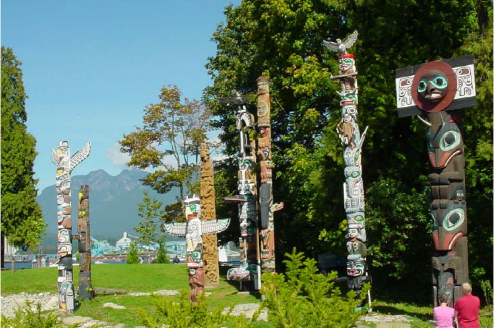Several intricately carved totem poles stand on a sunny lawn in front of lush trees and mountains. Two people are observing the impressive artworks.